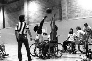 Two wheelchair basketball players going for the ball with a referee standing nearby and teammates ready in the background