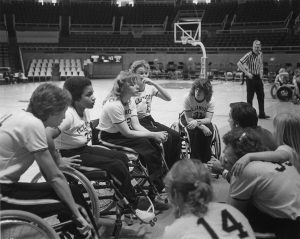 Women's Wheelchair basketball team in a huddle on sidelines with referee standing in the distance