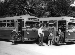 two buses with wheelchair lifts. Students in wheelchairs are using the bus and Nugent and another man stand next to the buses