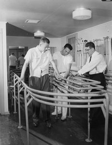 Man using curved rails to help him walk, while two other men look on