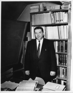 Professor Max Beberman standing in his office in front of bookshelves.