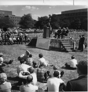 man on stage in front of harvested fields with people sitting in grass around stage