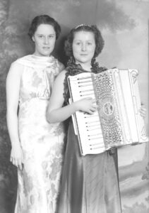 Two women in dresses, one is holding an accordian